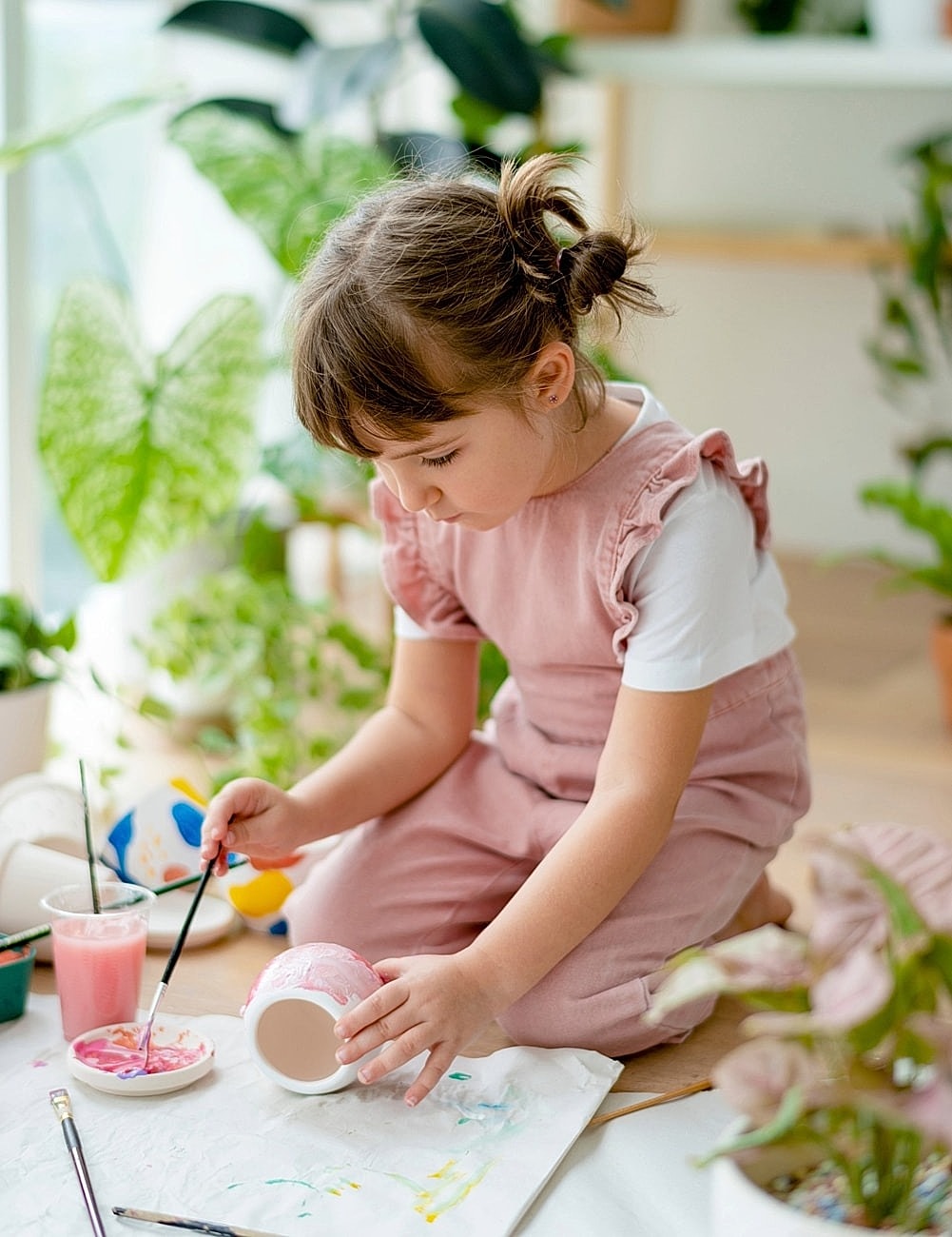 Child demonstrating fine motor strength through painting a pot among indoor plants.