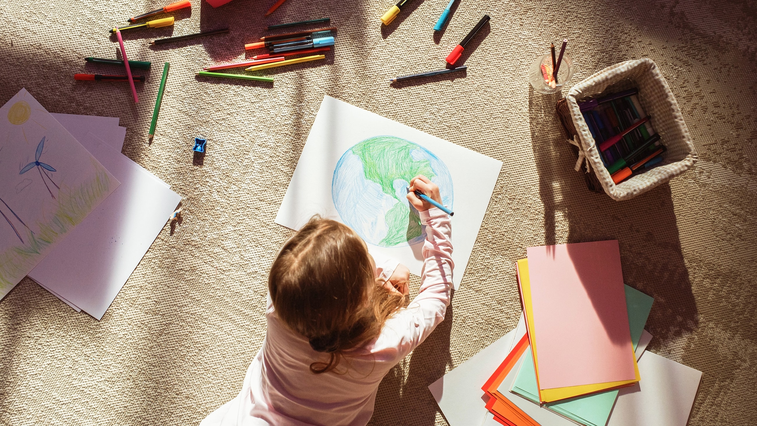 Child lying on floor, drawing a picture of planet Earth using colored pencils.